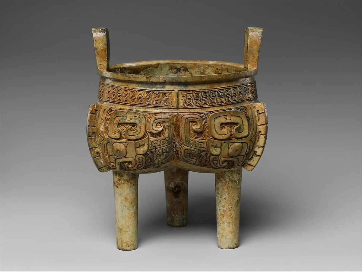 Shang bronze lobed tripod cauldron with three columnar legs, two upright handles, patterned bands and a mottled surface, photographed against gray.
